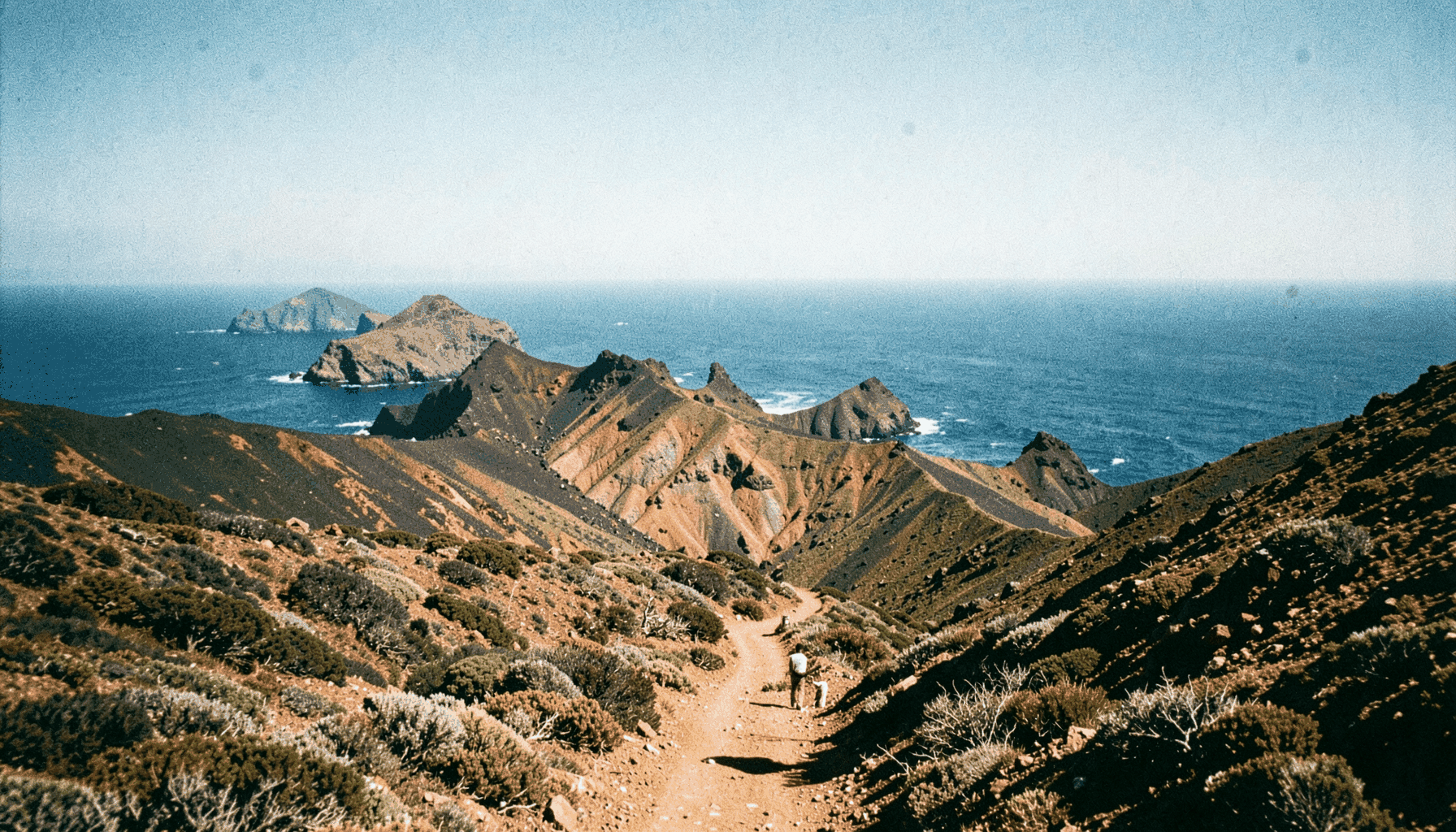 Sendero volcánico entre malpaíses y conos erosionados en la Isla de El Hierro, con el océano Atlántico al fondo bajo una luz intensa