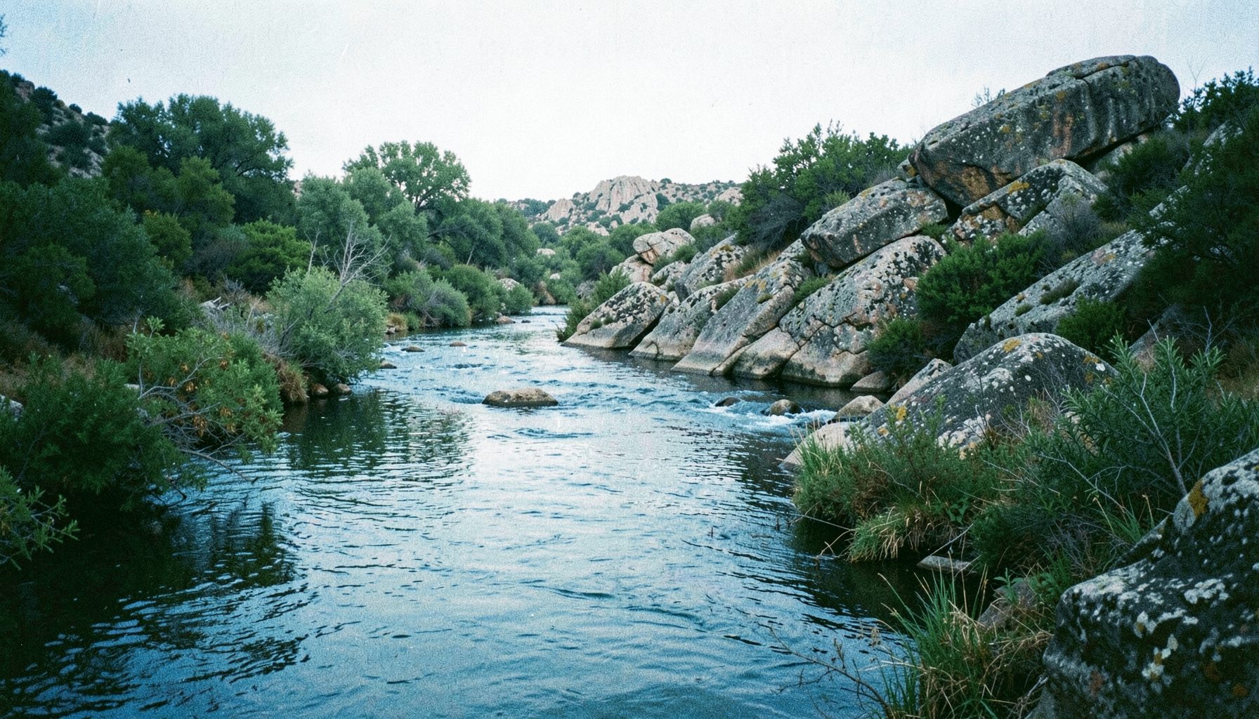 Senda junto al cauce del río Guadiana flanqueada por vegetación de ribera y grandes rocas graníticas, bajo una luz suave