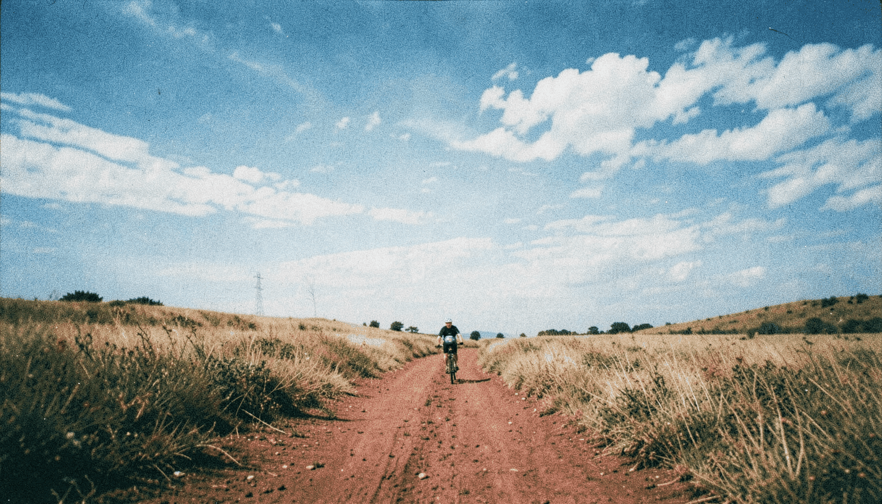 Fotografía analógica granulada de un ciclista en la lejanía ascendiendo por una pista de tierra rojiza entre pastizales y encinas