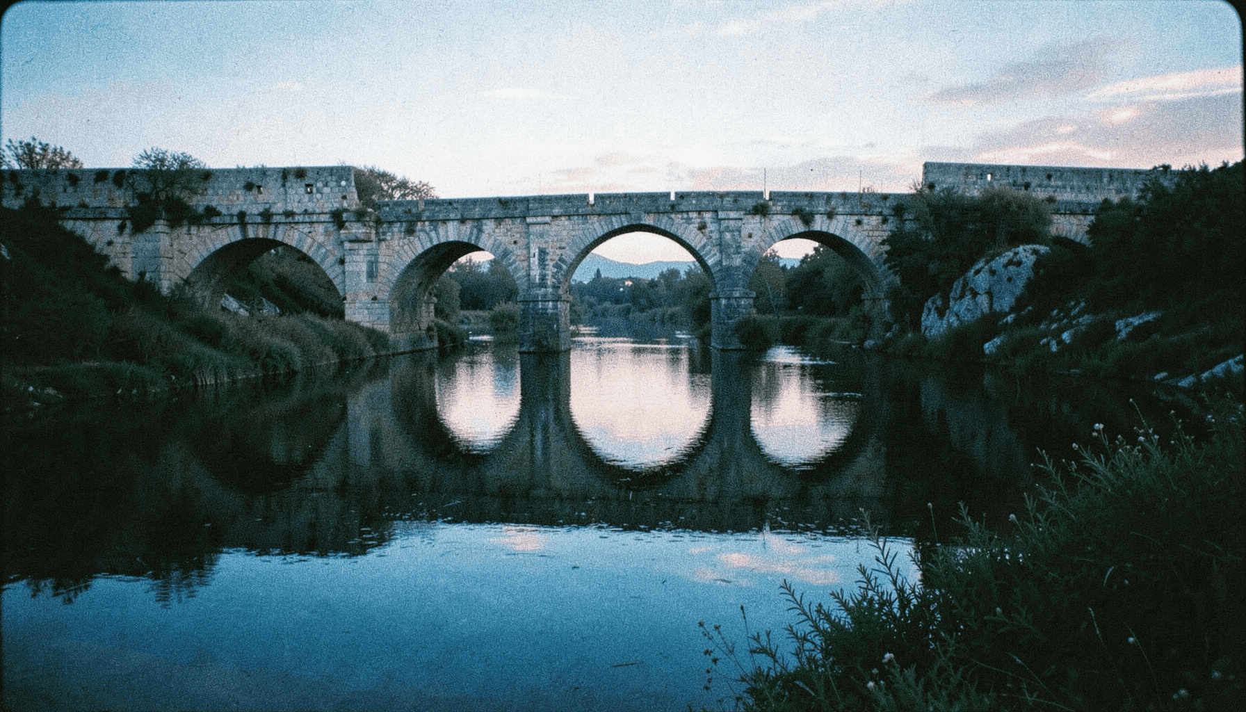 Puente romano de piedra sobre un río tranquilo reflejando el cielo del atardecer, vegetación de ribera, fotografía analógica