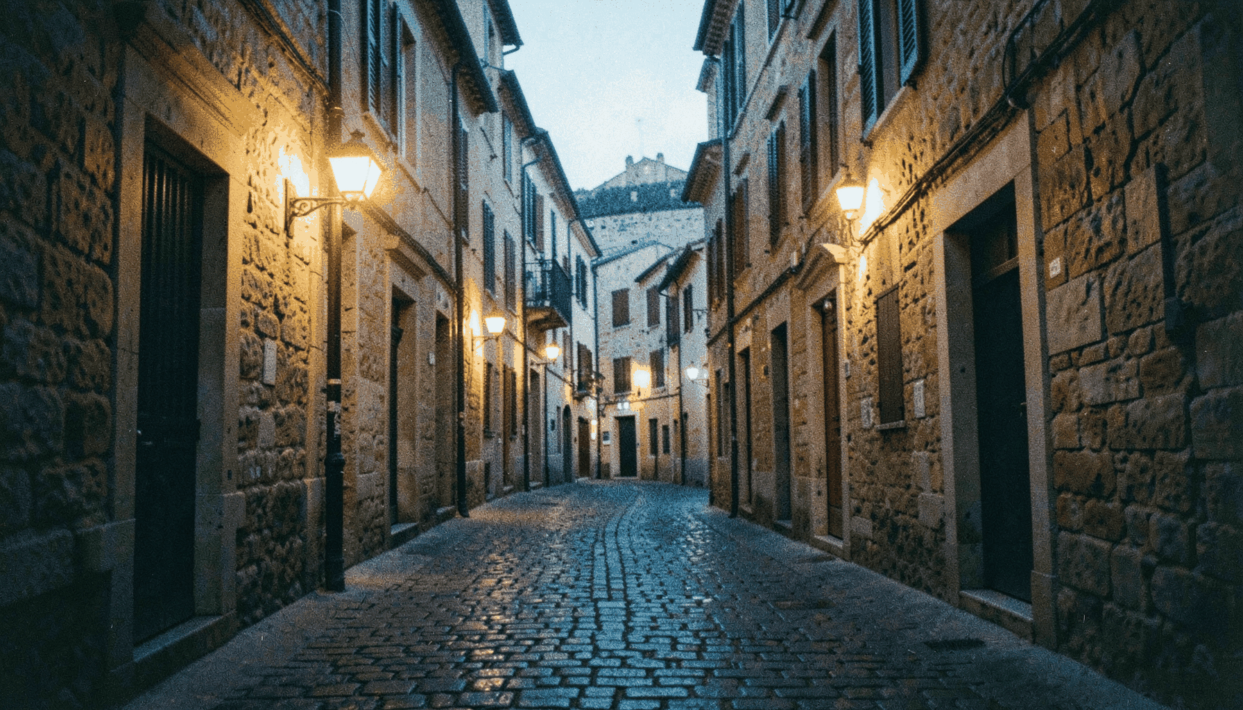 Calle empedrada de un casco histórico al atardecer, con fachadas de piedra dorada y farolas encendidas, atmósfera cálida