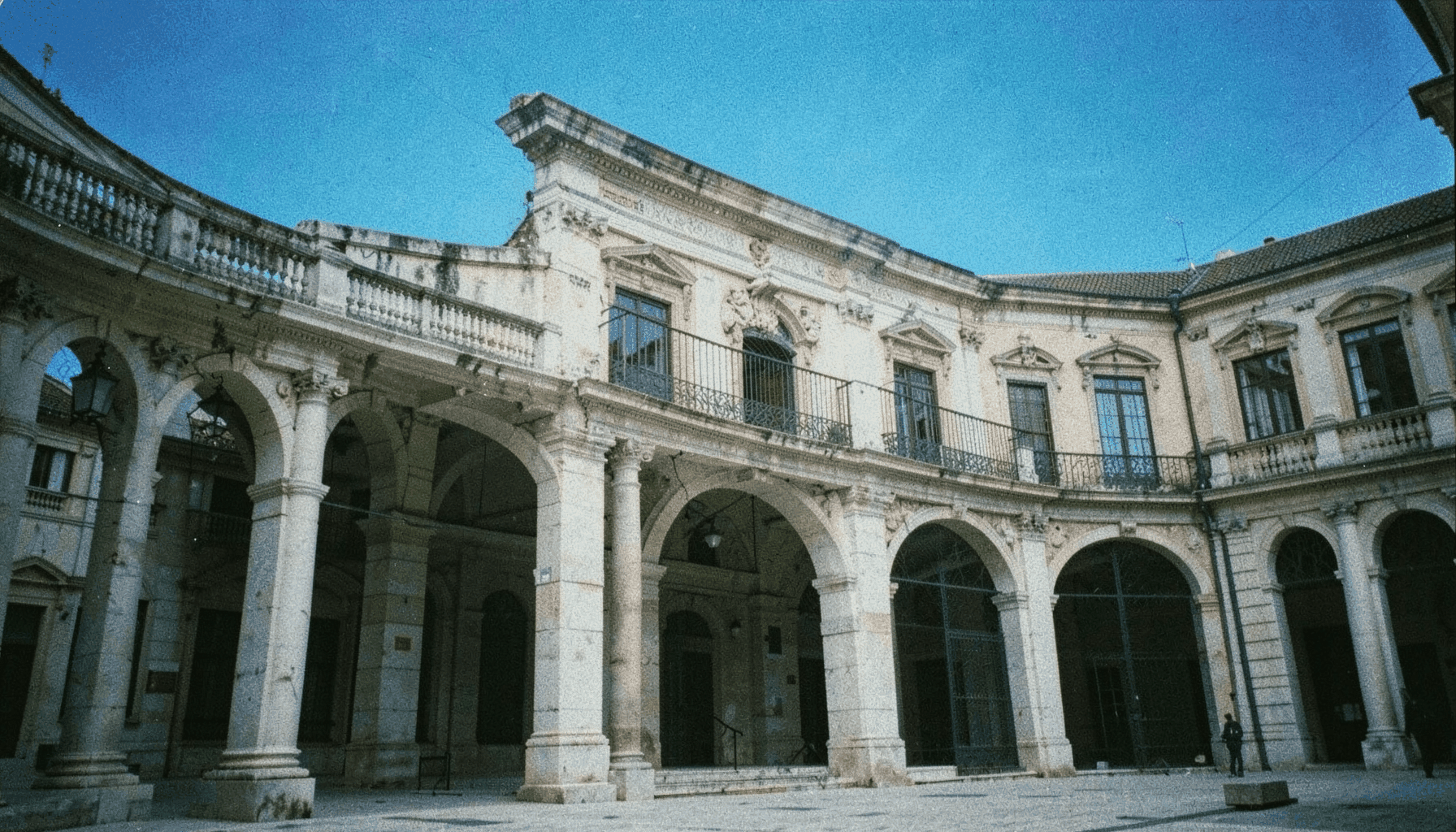 Plaza mayor porticada con soportales de granito y balcones de hierro forjado bajo un cielo azul intenso
