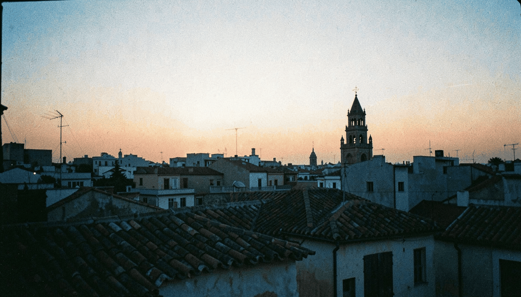Mirador urbano al atardecer con vistas a tejados de teja árabe y una torre campanario recortada contra el cielo anaranjado